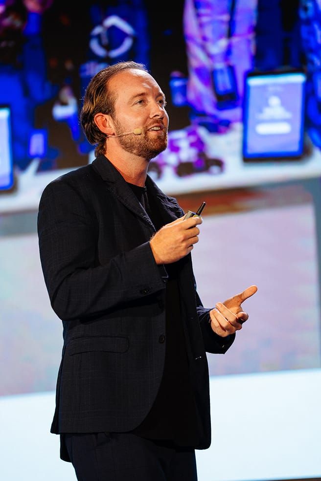 A man in a dark suit jacket and black shirt with a lapel microphone talks, holding a clicker.