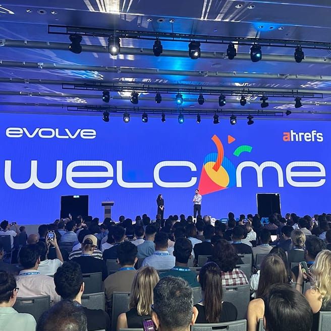 A conference room with a large screen displaying "evolve WELCOME ahrefs" and a colorful abstract design. Two speakers stand on stage, addressing a large audience seated in rows.