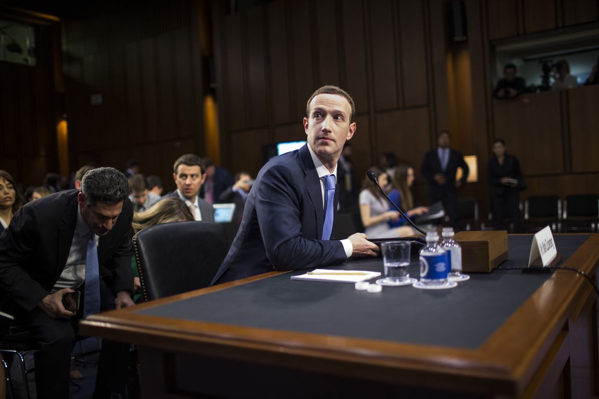 Mark Zuckerberg sitting at a hearing with reporters and onlookers seated behind him.