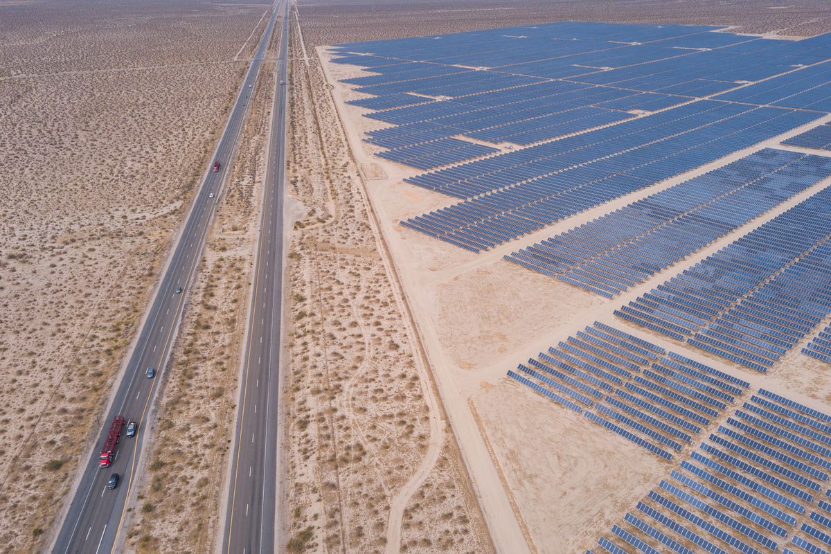 A bird’s-eye view of a vast solar farm in the Mojave desert in California shows hundreds of solar panels laid out in neat rows. The trucks and cars on a highway running parallel to the solar farm look tiny by comparison.