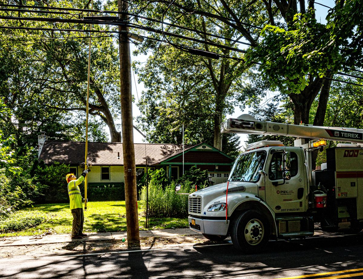 A utility worker standing on a tree-shaded residential street beside a utility truck uses a long yellow pole to adjust a power line high above them.