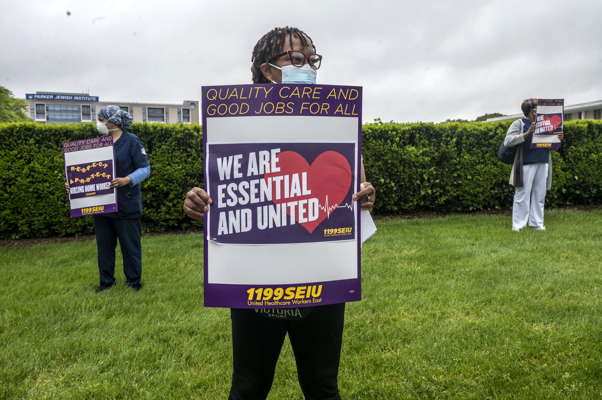 Health care workers outside a care facility hold signs that read, “Quality care and good jobs for all. We are essential and united. 1199 SEIU.”