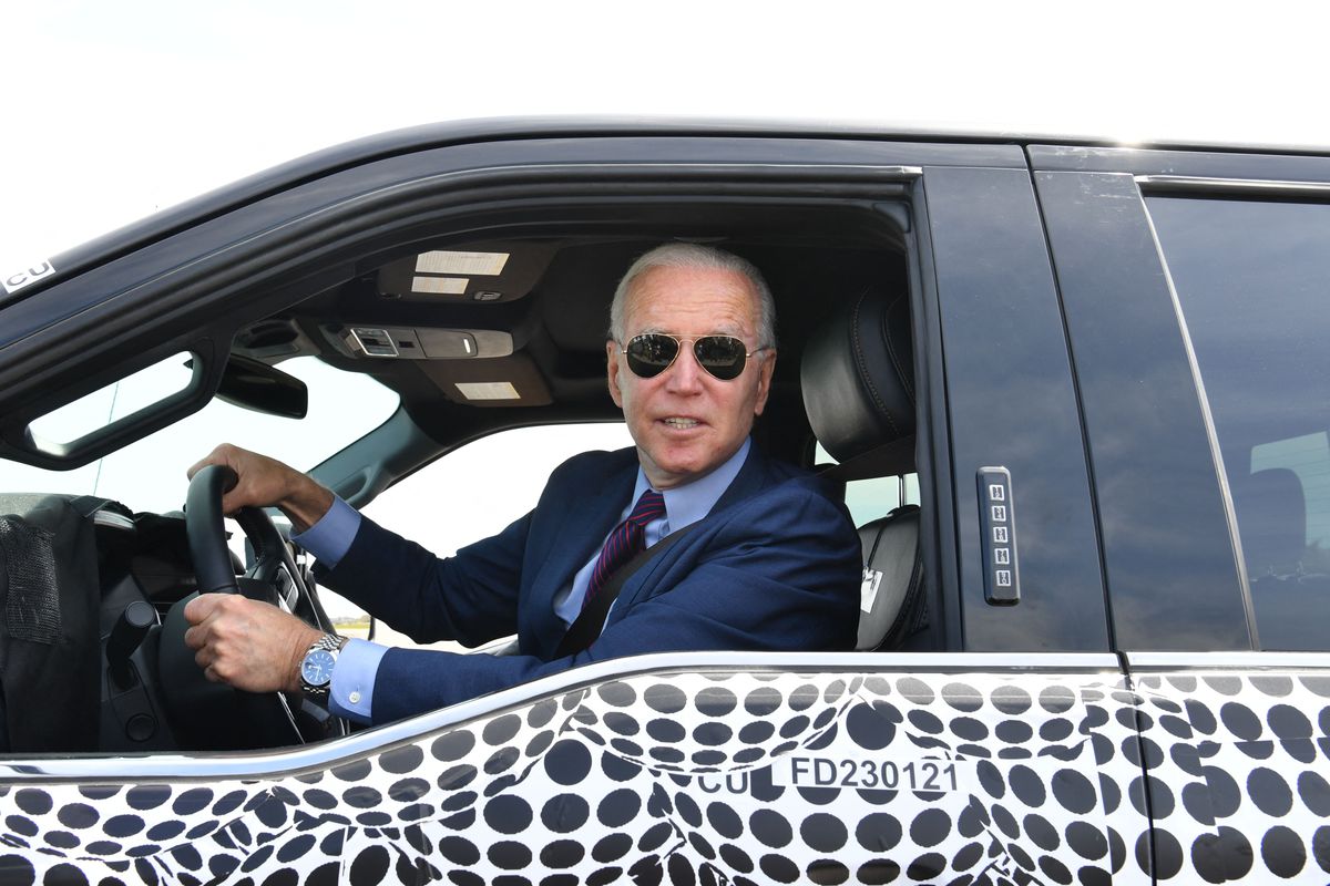 President Joe Biden, wearing his signature aviator sunglasses and with his hands on the steering wheel, looks at news photographers from the driver’s seat of an F-150 Lightning pickup truck.