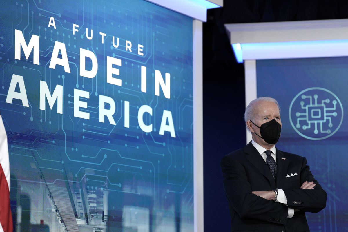 President Biden stands with arms crossed and face masked in front of a sign that reads “A future made in America.”