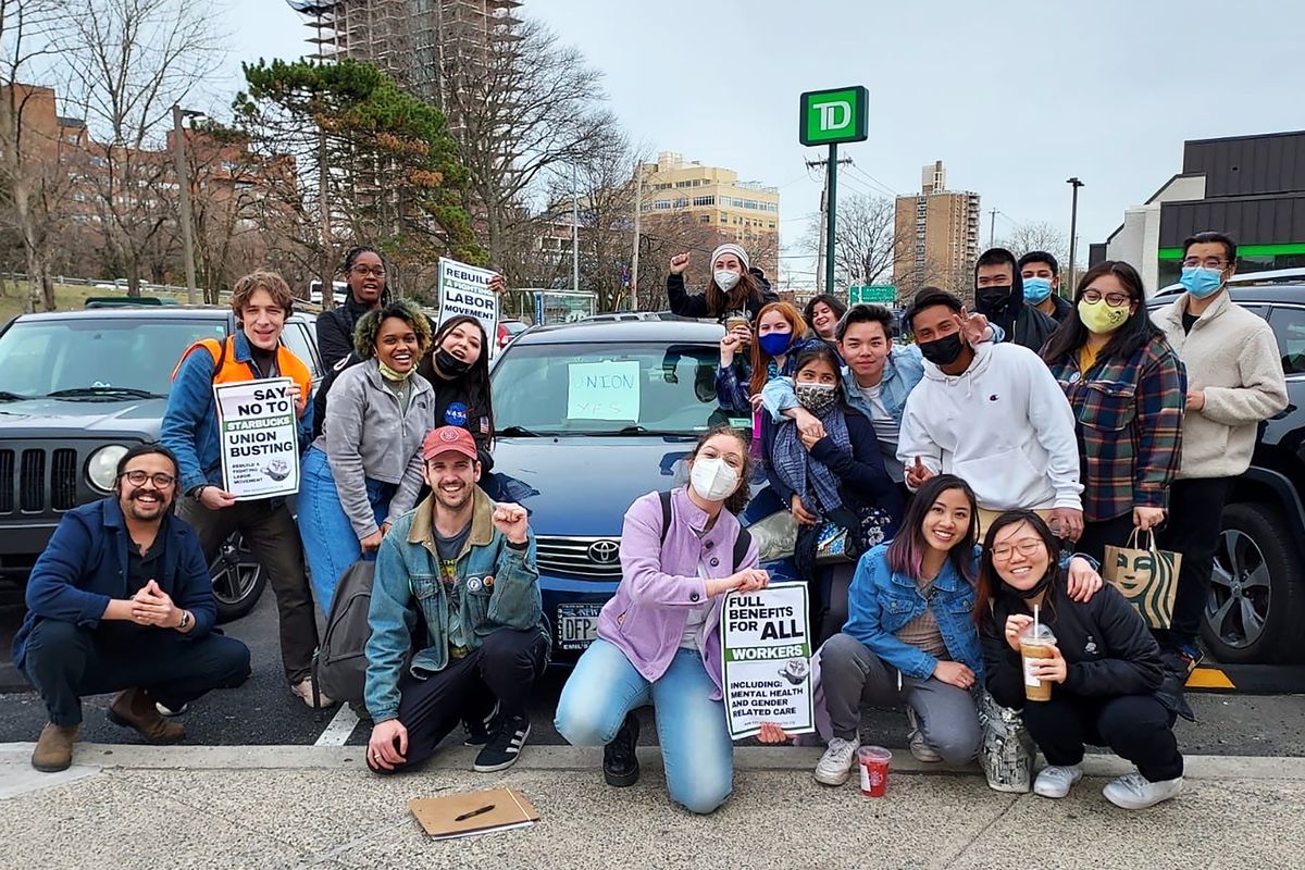 Members of Socialist Alternative NYC gather for a group photo around a car in a parking lot while holding signs in support of unions and workers.