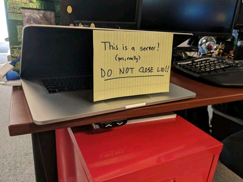 A photo of a laptop that is half-closed with a sheet of paper from a yellow legal pad that says This 9s a server (yes, really), do not close the lid!