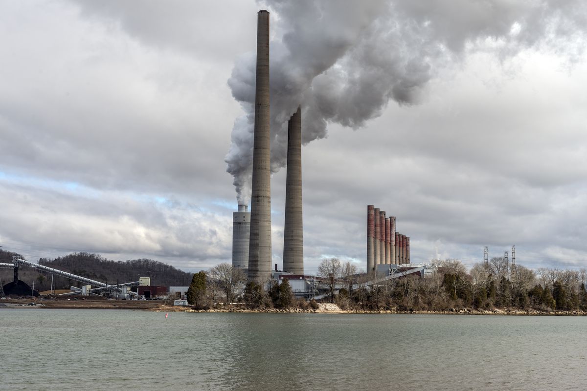 Black smoke rises from smokestacks at a power plant in front of a steely-colored body of water.