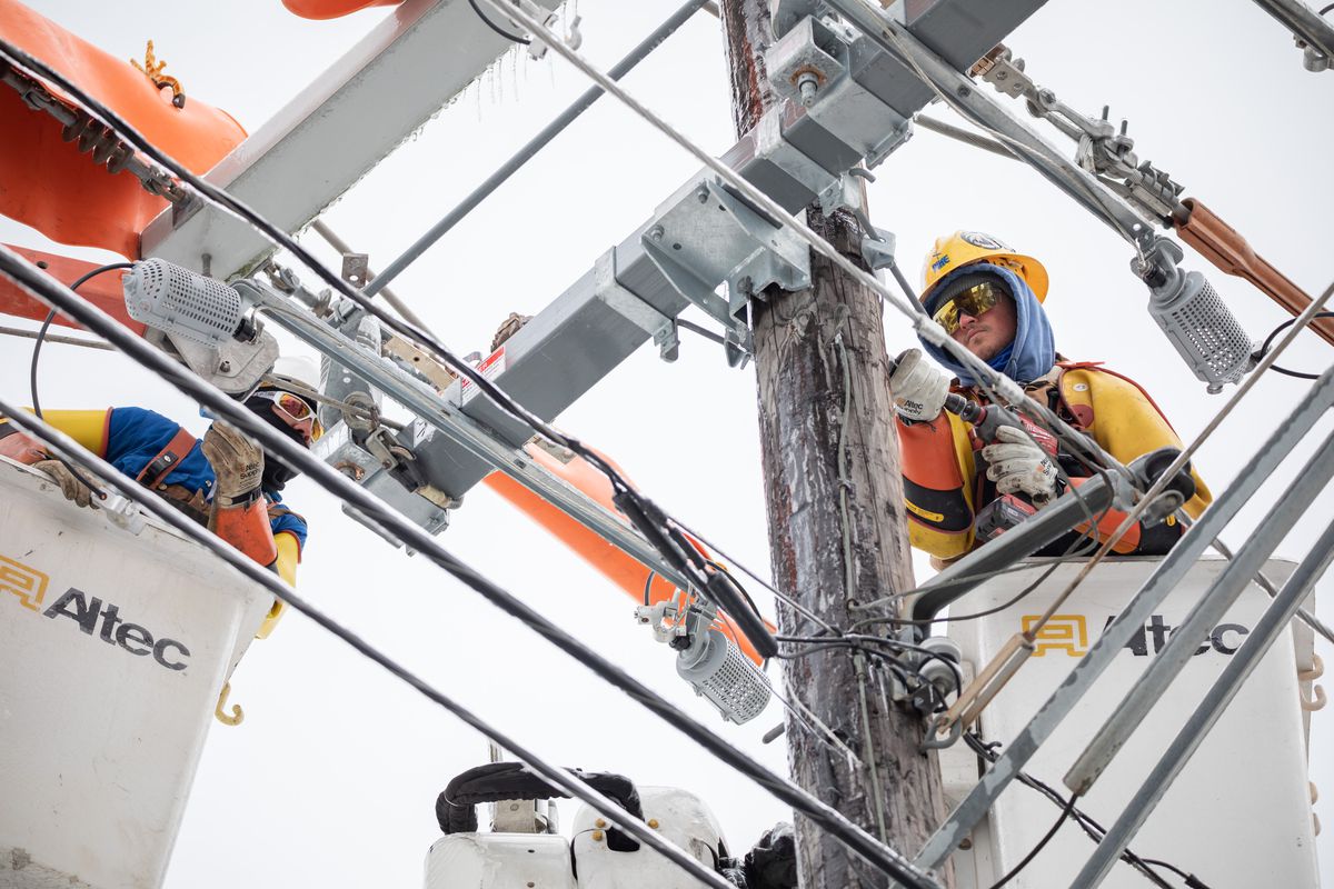 Utility workers in cherry-picker baskets wear winter gear and hard hats to work high up on a power pole coated in frost and icicles.